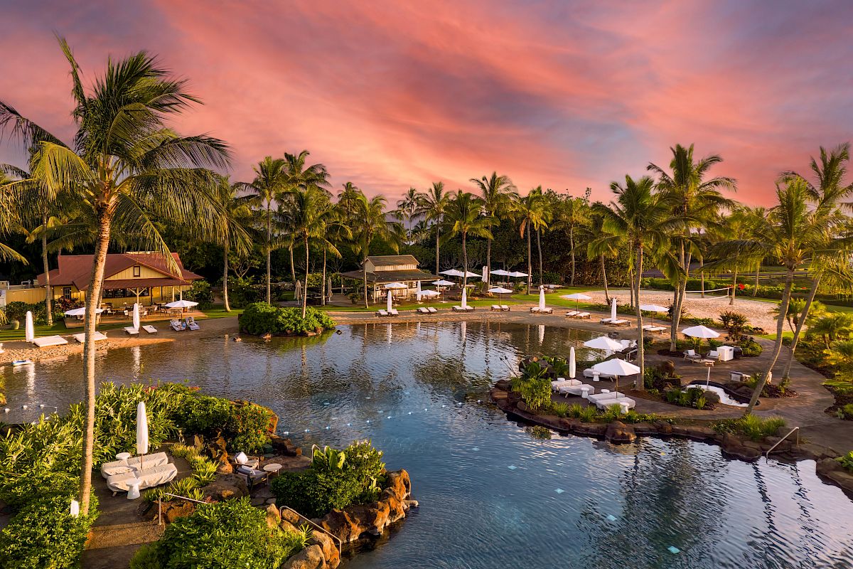 A tropical resort with palm trees, a lagoon-style pool, loungers, and a sunset-lit sky, creating a serene and picturesque setting.
