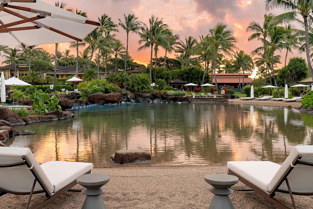Two lounge chairs face a serene pool surrounded by lush greenery, palm trees, and umbrellas at sunset, creating a peaceful tropical resort setting.