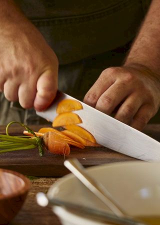 A person is chopping carrots on a wooden cutting board. Bowls and greenery are visible nearby.