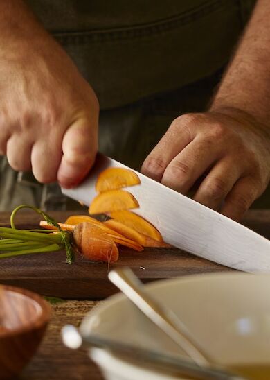 A person is chopping carrots on a wooden cutting board. Bowls and greenery are visible nearby.