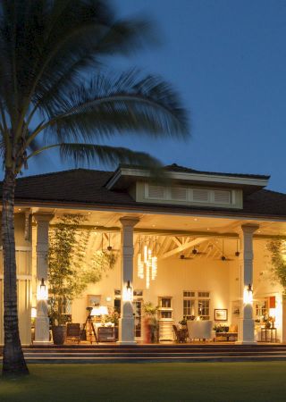 A large, illuminated house with open walls, surrounded by palm trees and lush greenery, set against an evening sky backdrop.