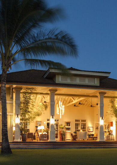 A large, illuminated house with open walls, surrounded by palm trees and lush greenery, set against an evening sky backdrop.