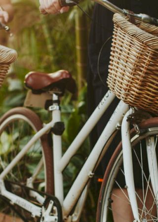 Two people are holding bicycles with wicker baskets on the handlebars, surrounded by lush greenery.