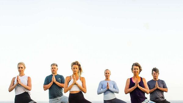 A group of six people is practicing yoga outdoors, performing tree pose on yoga mats with an ocean in the background.