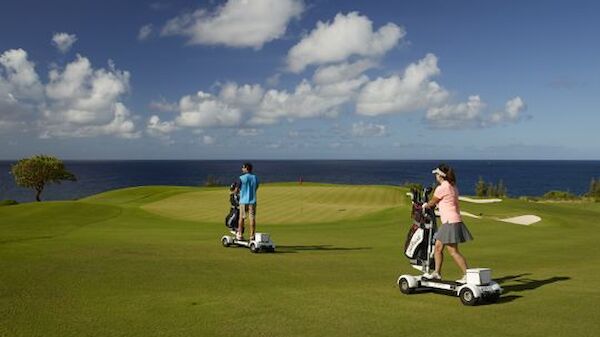 Two people are riding golf scooters on a golf course near the ocean under a blue sky with scattered clouds.