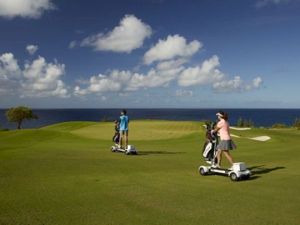 Two people are riding golf scooters on a golf course near the ocean under a blue sky with scattered clouds.