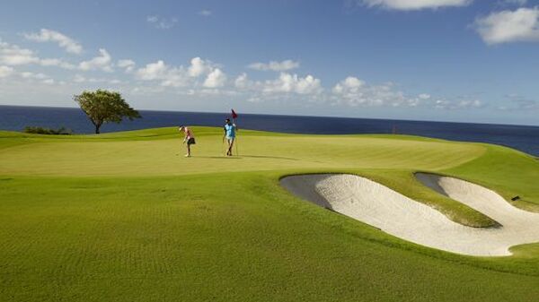 Two people playing golf on a green course near the ocean, with a sand bunker and bright blue sky with scattered clouds.