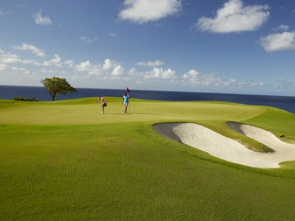 Two people playing golf on a green course near the ocean, with a sand bunker and bright blue sky with scattered clouds.