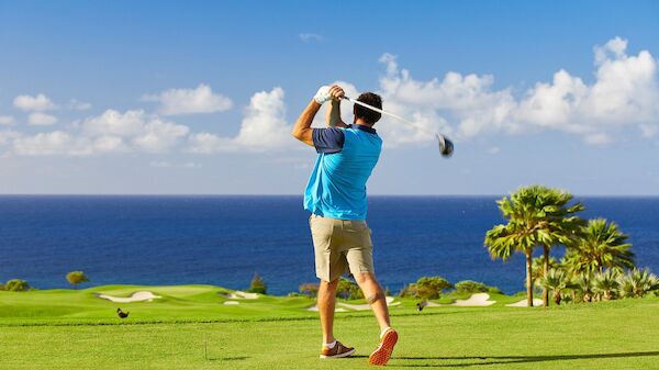 A person is playing golf on a lush green course with an ocean view in the background, under a bright blue sky with scattered clouds.