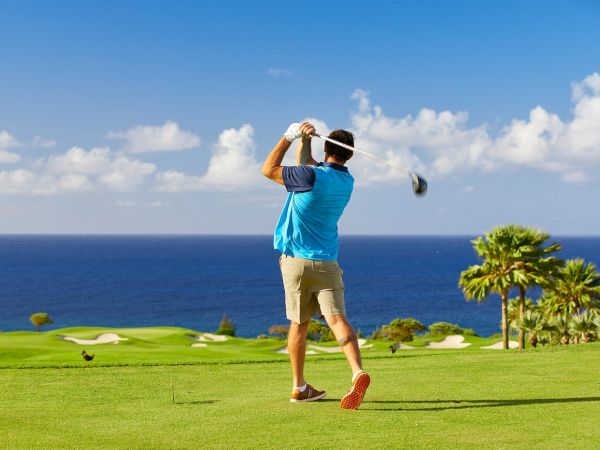 A person is playing golf on a lush green course with an ocean view in the background, under a bright blue sky with scattered clouds.