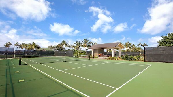 A tennis court with green surface, white lines, a net, surrounded by fencing. There is a small pavilion and palm trees under a blue sky with clouds.