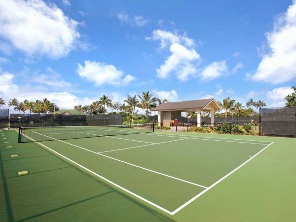 A tennis court with green surface, white lines, a net, surrounded by fencing. There is a small pavilion and palm trees under a blue sky with clouds.