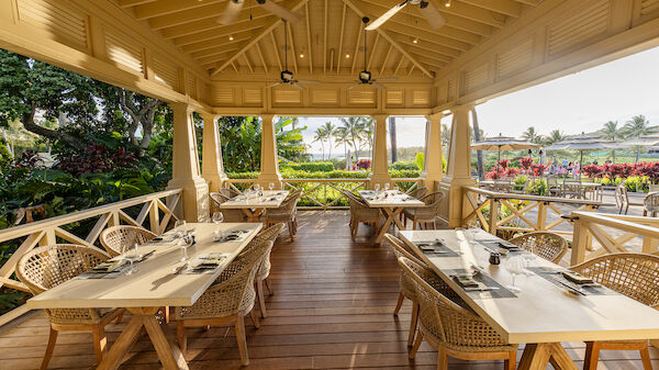 The image shows an outdoor dining area with wooden tables, rattan chairs, and tropical plants, creating a serene atmosphere.
