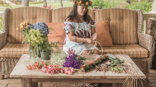 A person on a sofa is making a floral wreath. The table in front has flowers, leaves, and a jar of hydrangeas. It’s an outdoor setting, an open green area.