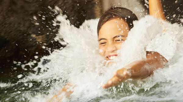 A person is joyfully splashing in the water, likely at a pool or water park, with water spraying around them as they smile.