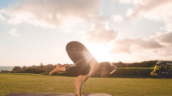 A person is performing a yoga pose on a mat outdoors with the sun setting in the background, creating a serene atmosphere.