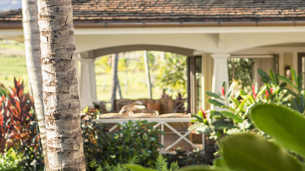 A beautiful porch area of a house with surrounding greenery, palm tree trunks, and distant scenery visible through an archway in the background.