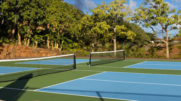 The image shows a tennis court with blue and green surfaces surrounded by trees, under a clear sky with a few clouds, and a wooden bench is in the foreground.