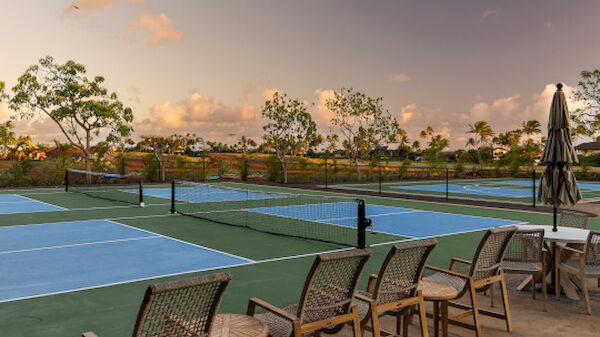 The image shows outdoor tennis courts with blue surfaces and green surroundings, several chairs, a table, and an umbrella beside the court.