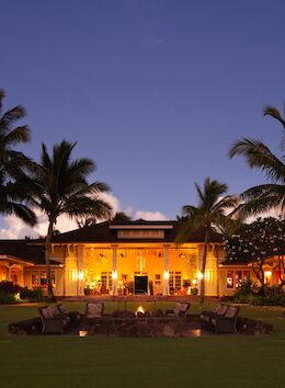 A beautifully lit tropical resort at dusk, featuring palm trees, a central building with warm lights, and an outdoor seating area with a fire pit.