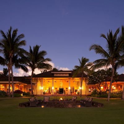 A beautifully lit tropical resort at dusk, featuring palm trees, a central building with warm lights, and an outdoor seating area with a fire pit.