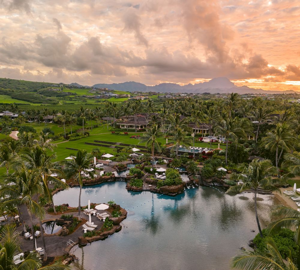 A tropical resort with a large pool, lush greenery, palm trees, and mountains in the background during a sunset with a cloudy sky.