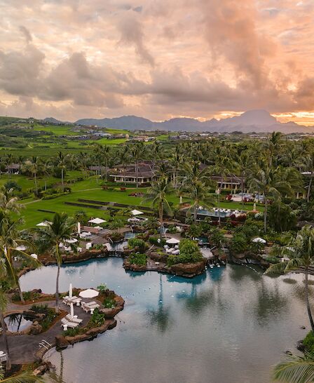 A tropical resort with a large pool, lush greenery, palm trees, and mountains in the background during a sunset with a cloudy sky.