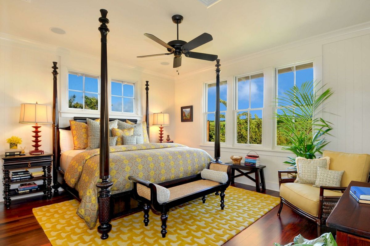 A bright bedroom with a four-poster bed, yellow and gray bedding, two nightstands with lamps, a ceiling fan, and large windows letting in natural light.