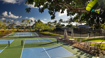 The image shows a sports facility with tennis and pickleball courts, surrounded by greenery, palm trees, and outdoor seating under an umbrella.