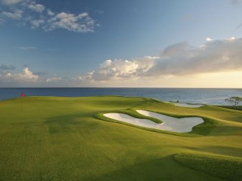 A scenic golf course by the ocean, featuring a well-maintained green, a sand bunker, and a red flag marking the hole.