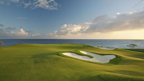 A scenic golf course by the ocean, featuring a well-maintained green, a sand bunker, and a red flag marking the hole.