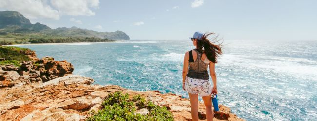 A woman with a hat stands on a rocky coastline, overlooking the ocean. The scene includes cliffs, greenery, and mountains in the background.