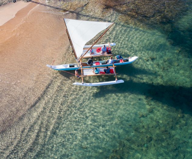 An aerial view shows a sailboat with people on board near a sandy beach and clear, shallow waters, casting a shadow on the ocean floor.