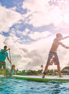 Three people are paddleboarding on a sunny day, with palm trees and a bright sky in the background.