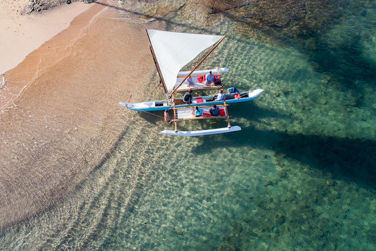 A small boat with a sail and several passengers is docked at a sandy shore, with clear, shallow water surrounding it.