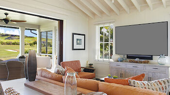 A cozy living room with a ceiling fan, a large TV, beige couches, and a view of a green landscape through an open sliding door.