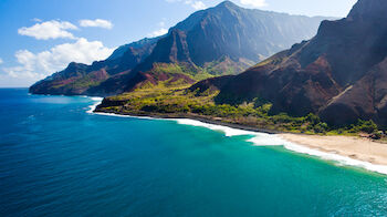 A stunning coastal view with clear blue water, a sandy beach, and steep, lush green mountains under a bright sky.