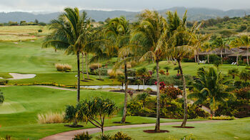 A scenic view of a lush golf course with palm trees, a paved path, and rolling hills in the background under a cloudy sky.