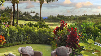 An outdoor seating area with wicker chairs around a fire pit, surrounded by lush greenery, colorful plants, and a scenic view of palm trees and fields.