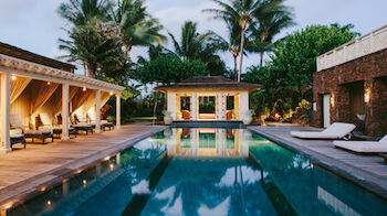 A luxurious outdoor pool area with lounge chairs, cabanas, palm trees, and a modern, well-lit pool house in the background.