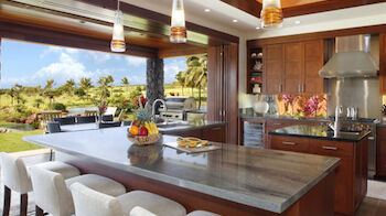 This image shows a modern kitchen with wooden cabinets, an island with bar stools, pendant lighting, and a view of an outdoor patio and garden.