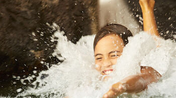 A person is joyfully splashing into a pool from a water slide, with water spraying around them and their feet and arms visible.