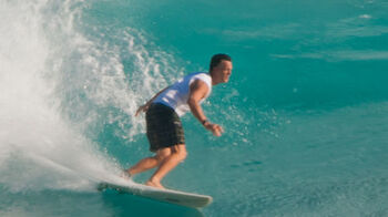 A person is surfing on a wave in the ocean, balancing on a surfboard while wearing a white shirt and black shorts.