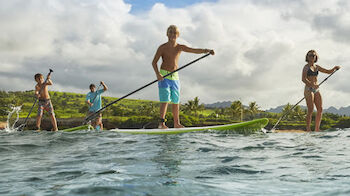 Four individuals are stand-up paddleboarding on a body of water, with greenery and a cloudy sky in the background.