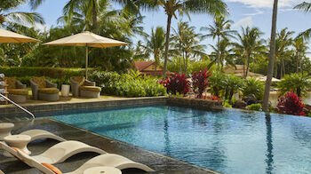 The image shows a luxurious outdoor pool area with white lounge chairs, umbrellas, tropical plants, and palm trees under a bright blue sky.