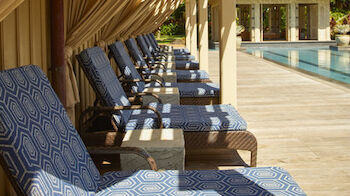 This image shows a row of poolside lounge chairs with blue patterned cushions under a series of tents, next to a swimming pool surrounded by palm trees.