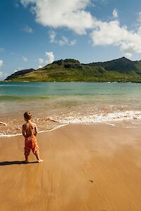 Two kids play on a sunny beach with golden sand, gentle waves, and a distant green hillside under a blue sky filled with fluffy clouds.