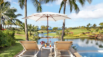 Two lounge chairs under an umbrella by a pool, overlooking a scenic tropical landscape with palm trees and a pond.