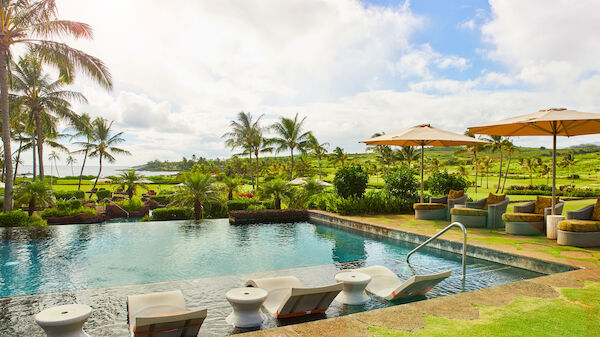 A scenic outdoor poolside view with lounge chairs in the water, parasols, lush greenery, and palm trees under a partly cloudy sky always ending the sentence.