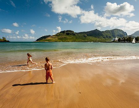 Two children are playing at the shore of a beach with clear blue water and lush green hills in the background, under a bright, partly cloudy sky.